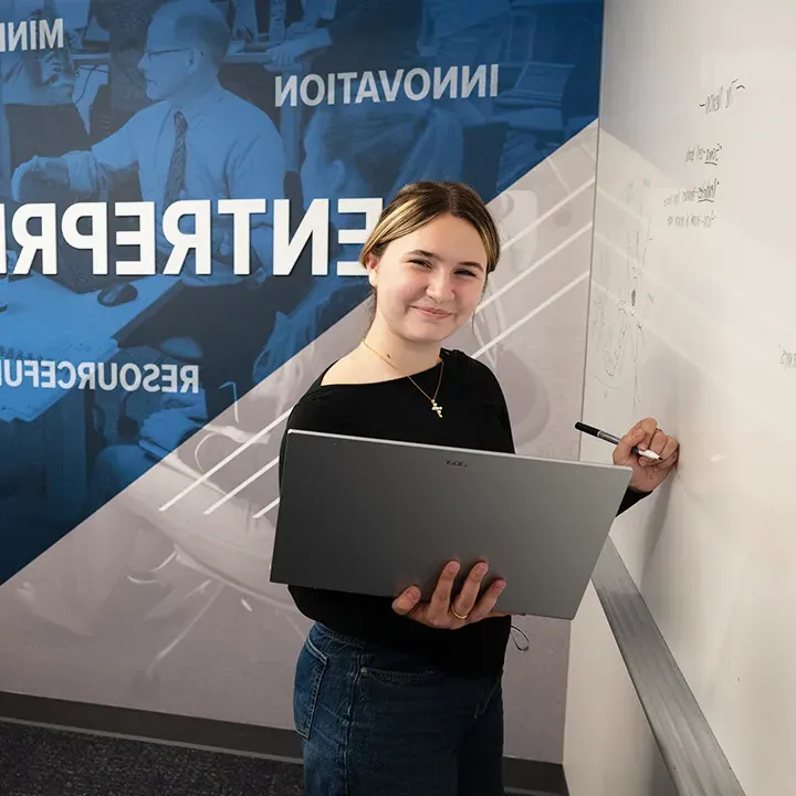 Student smiling while holding laptop in one hand and writing on whiteboard with the other hand.